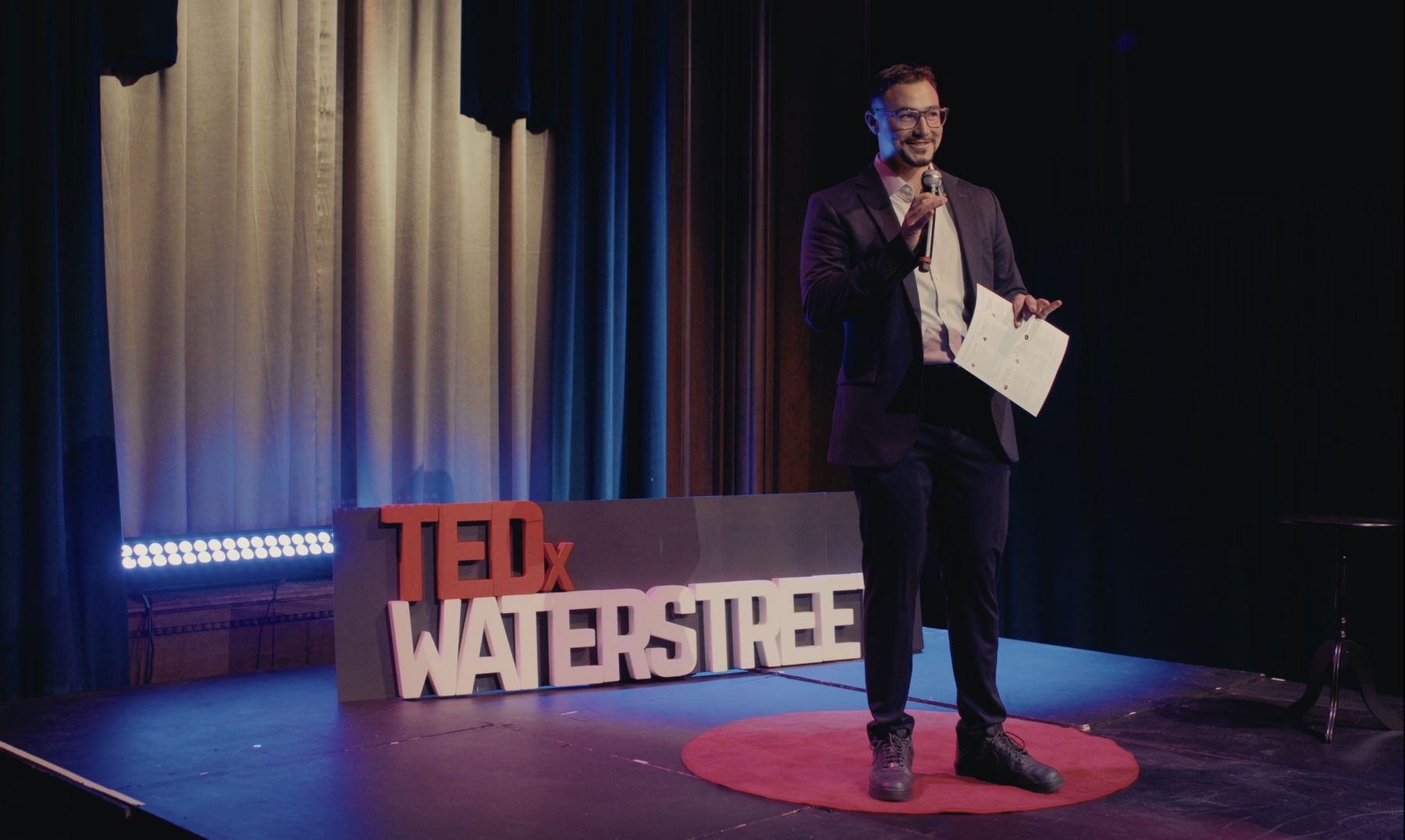 Dominic hosting TEDxWaterStreet beside the TEDx sign and red carpet